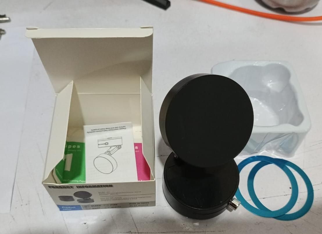 Black circular object next to an open box with product packaging on a white surface. Multifunctional phone vacuum holder with magnetic attachment showing retractable arm and 360 rotatable features